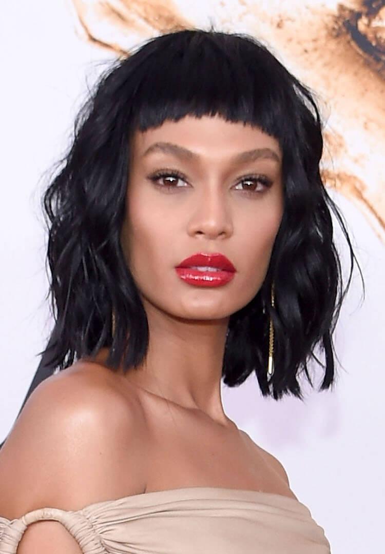 A photo of model Joan Smalls donning a chic nude off-shoulder dress with her striking black micro bangs wearing a bold swipe of vibrant red lipstick adorned with stylish bangles 