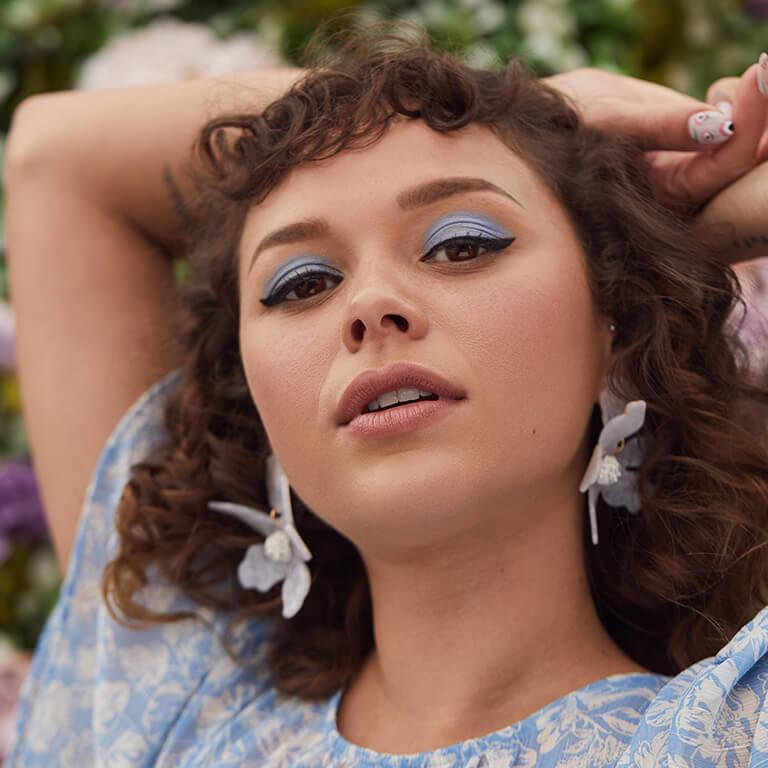 A close-up image of a model in a blue floral top rocking a light blue eyeshadow and black cat eyeliner look and wearing statement floral earrings