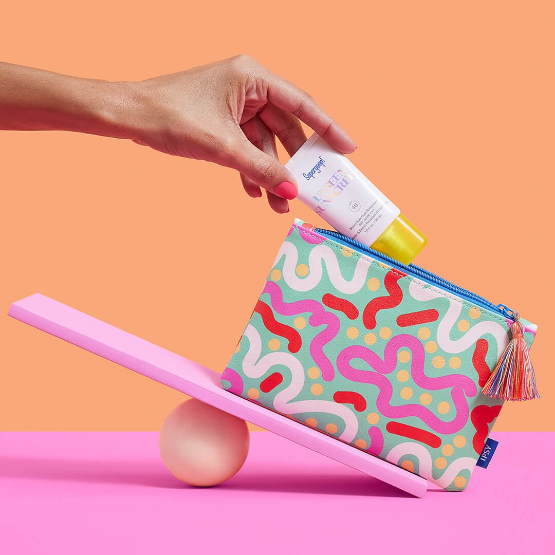 Woman's hand putting SUPERGOOP! inside a colorful makeup bag on a mini pink seesaw against orange and pink background
