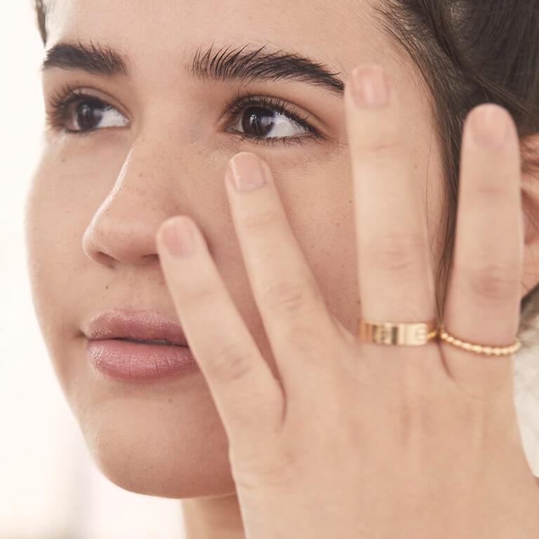 A closeup image of a model wearing gold rings applying concealer under eyes with her ring finger
