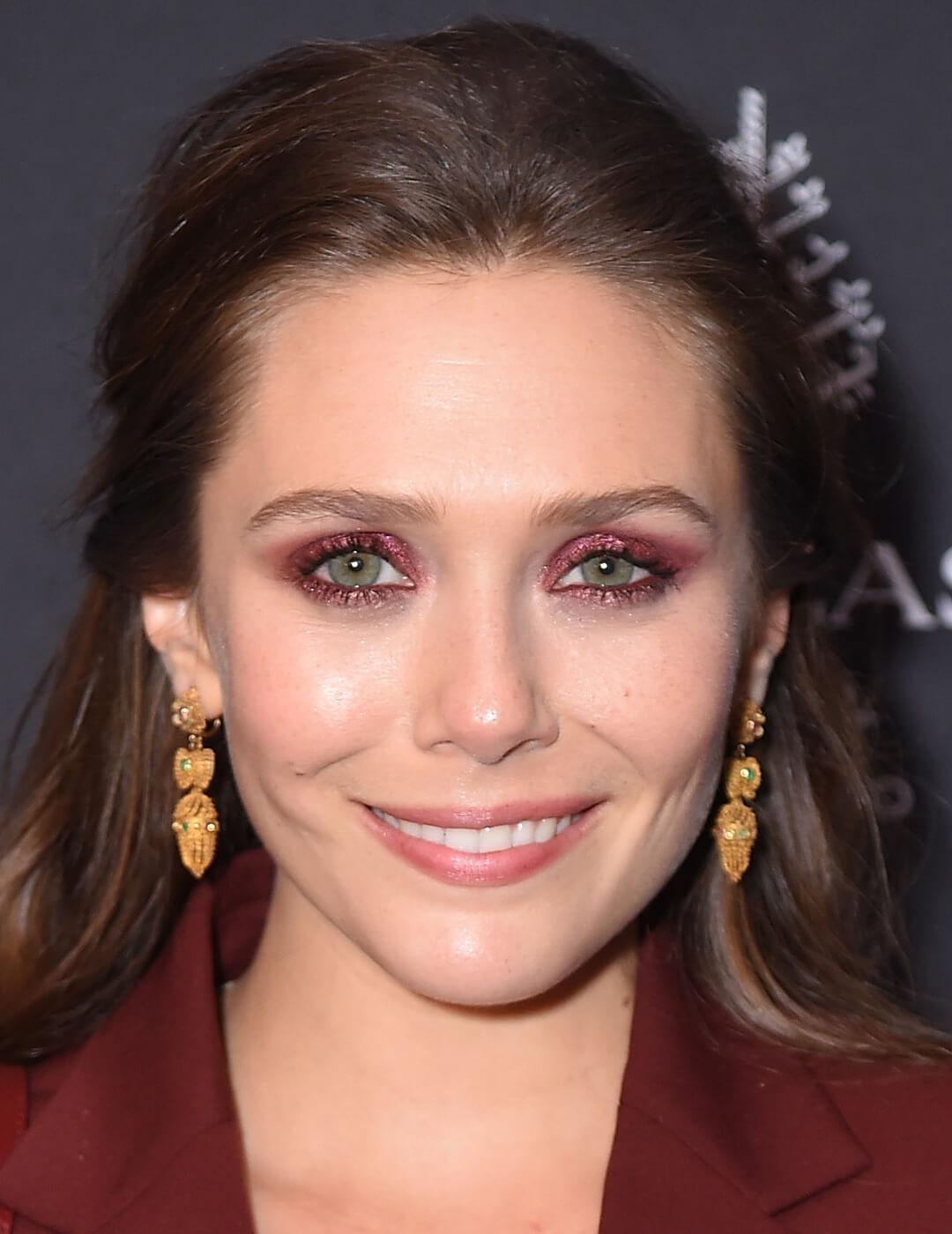 Close-up image of Elizabeth Olsen rocking a shimmery maroon smoky eye makeup look and gold dangling earrings
