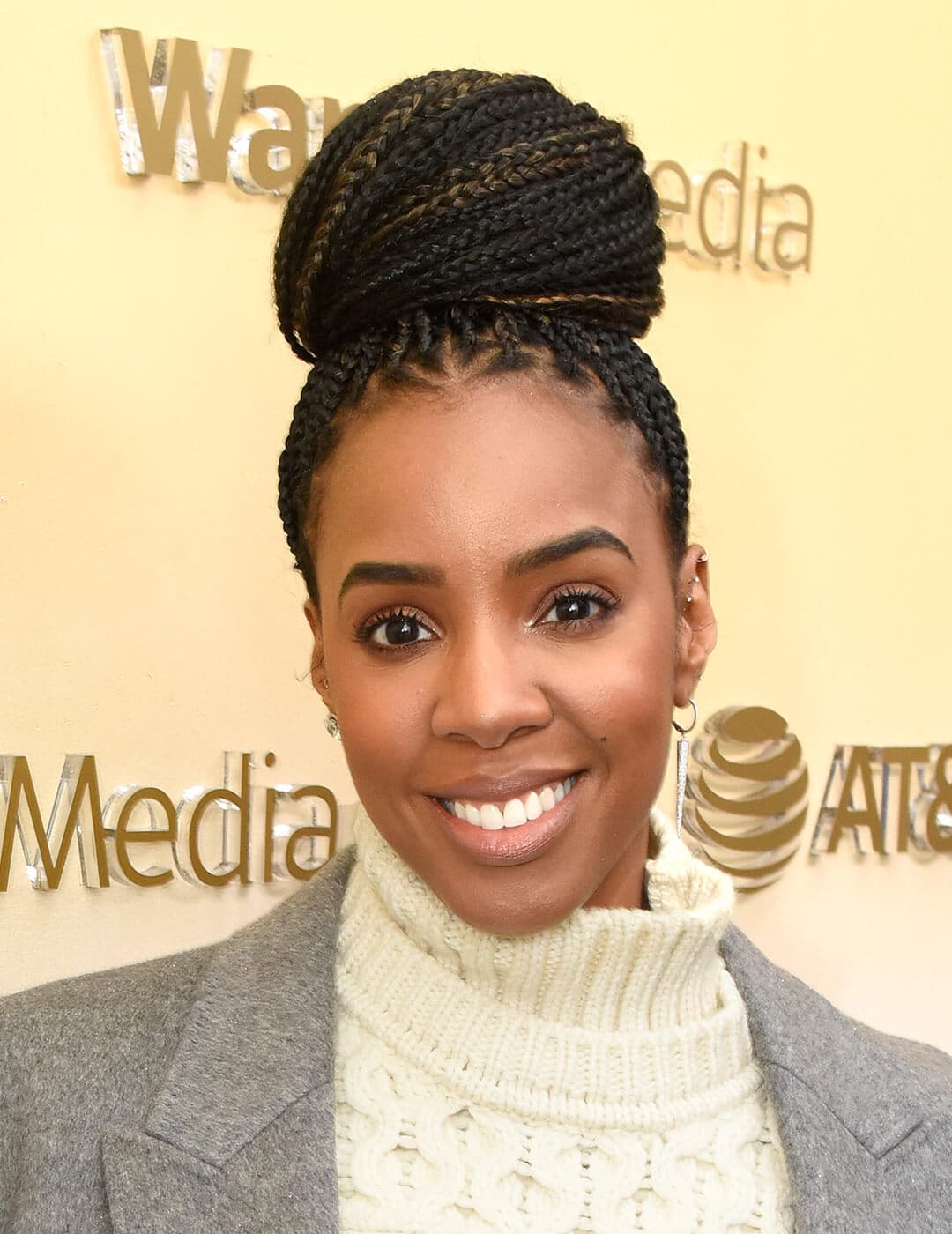 Kelly Rowland in an offwhite turtle neck sweater and grey coat rocking a braided top knot hairstyle