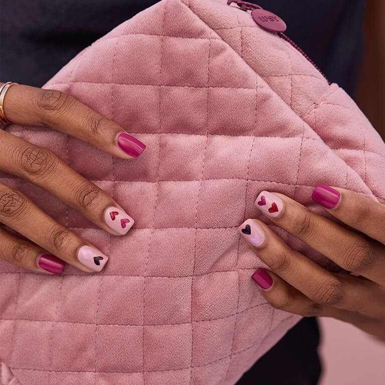 An image of two hands with light and dark pink hearts nail art holding a pink quilted bag