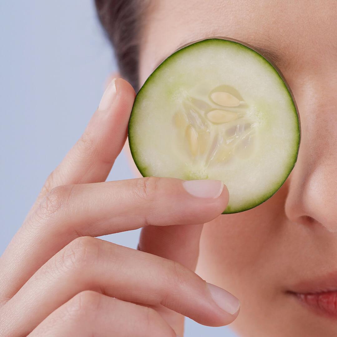 Close-up image of a woman holding a cucumber slice to cover her eye
