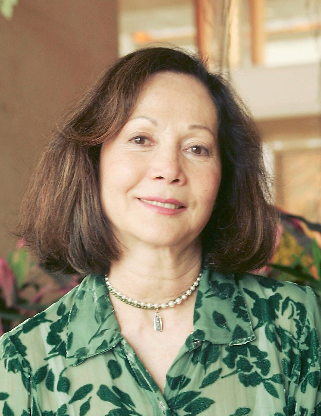 A photo of Nancy Kwan smiling in front of a camera, wearing a see through green floral long sleeve with her shoulder-length haircut