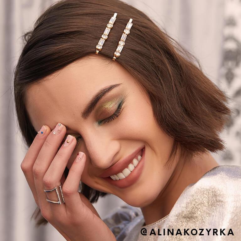 Model Alina Kozyrka wearing gemstone studded hairclips smiles while covering half of her face with her hand