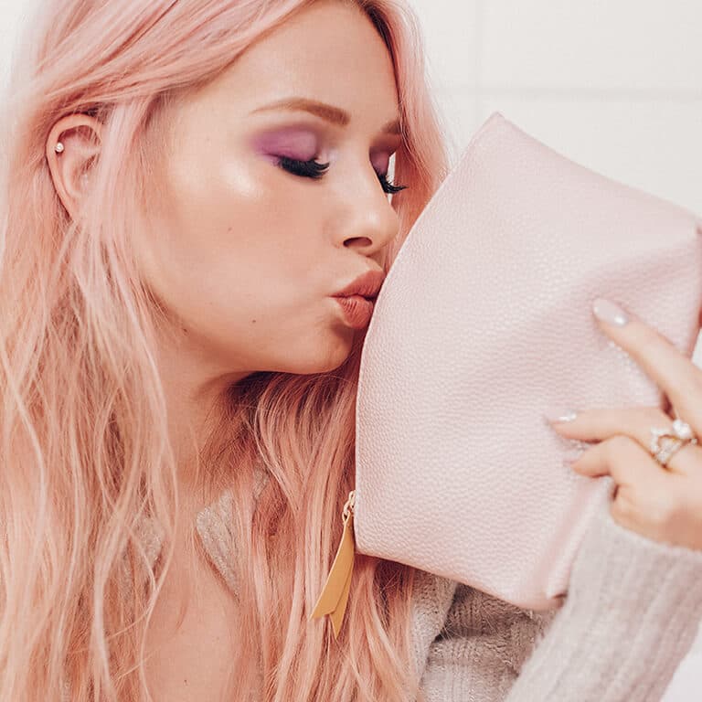 An image of a model with a purple and pink eyeshadow look kissing her soft pink makeup bag