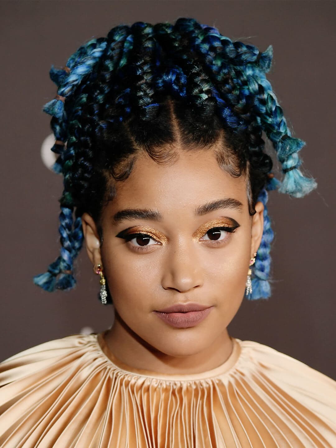 Amandla Stenberg in a gold, pleated dress rocking a black and gold eye makeup look and blue, braided high pony tail hairstyle