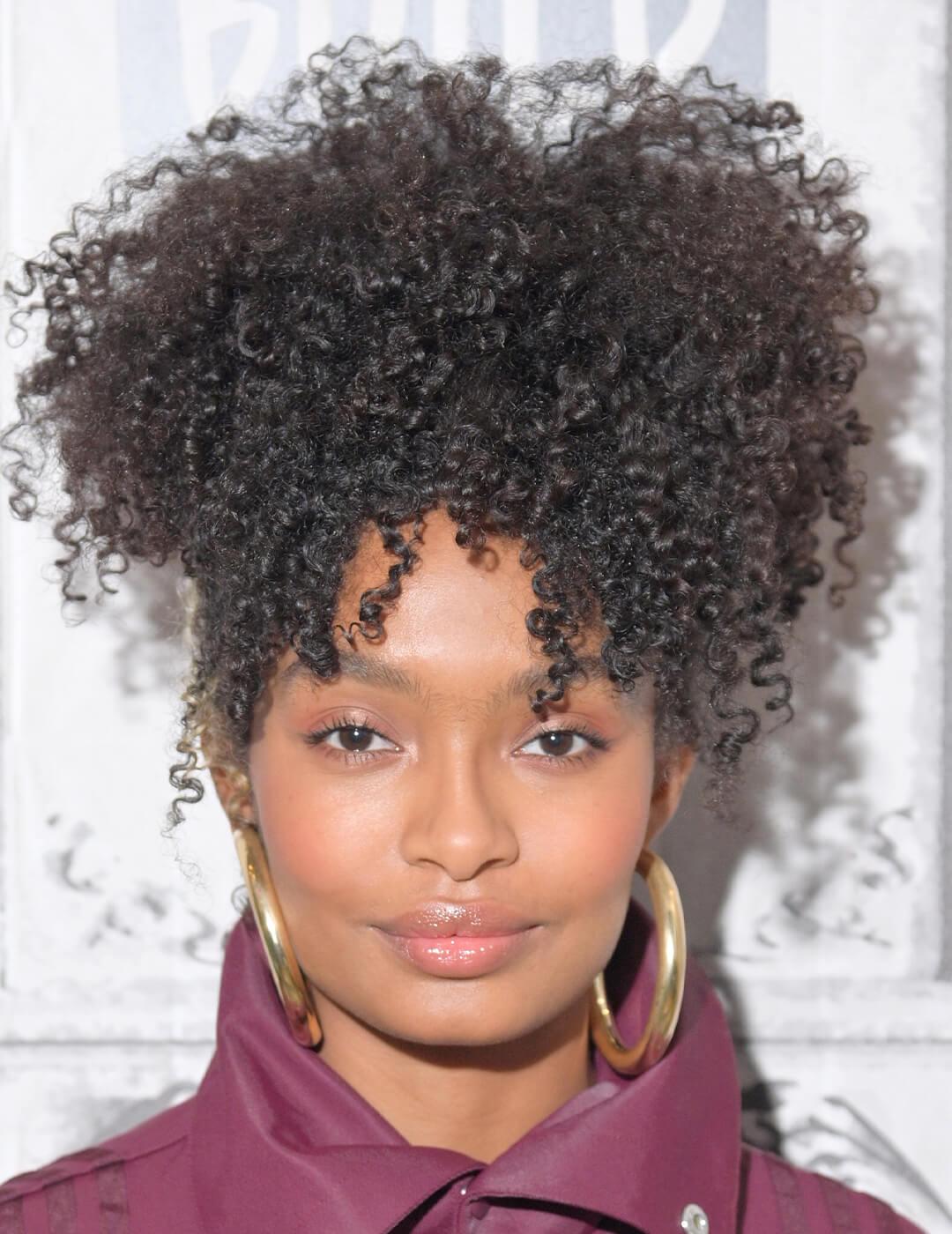 Close-up image of Yara Shahidi rocking a muted magenta coat, bold hoop earrings, and curly updo hairstyle