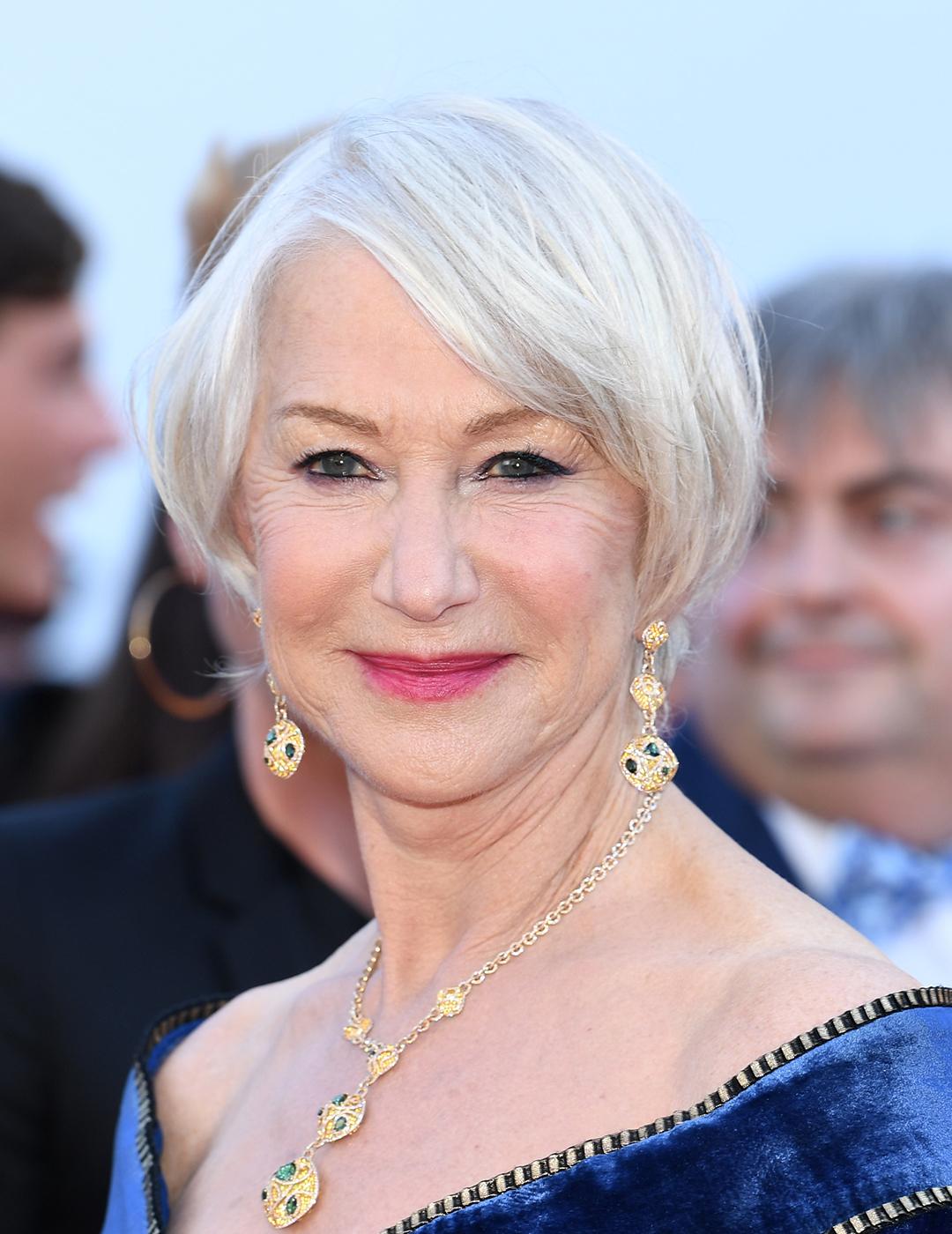 A photo of Helen Mirren wearing a blue velvet long dress paired with a dangling golden earrings and necklace perfect for her short bob hair