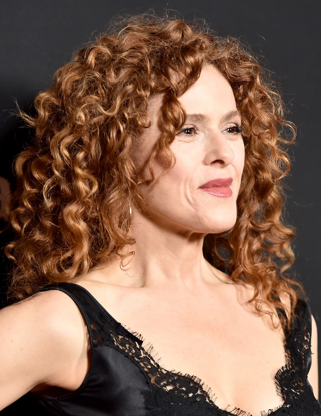 A photo of Bernadette Peters in her natural voluminous curls dressed in a black satin get up  