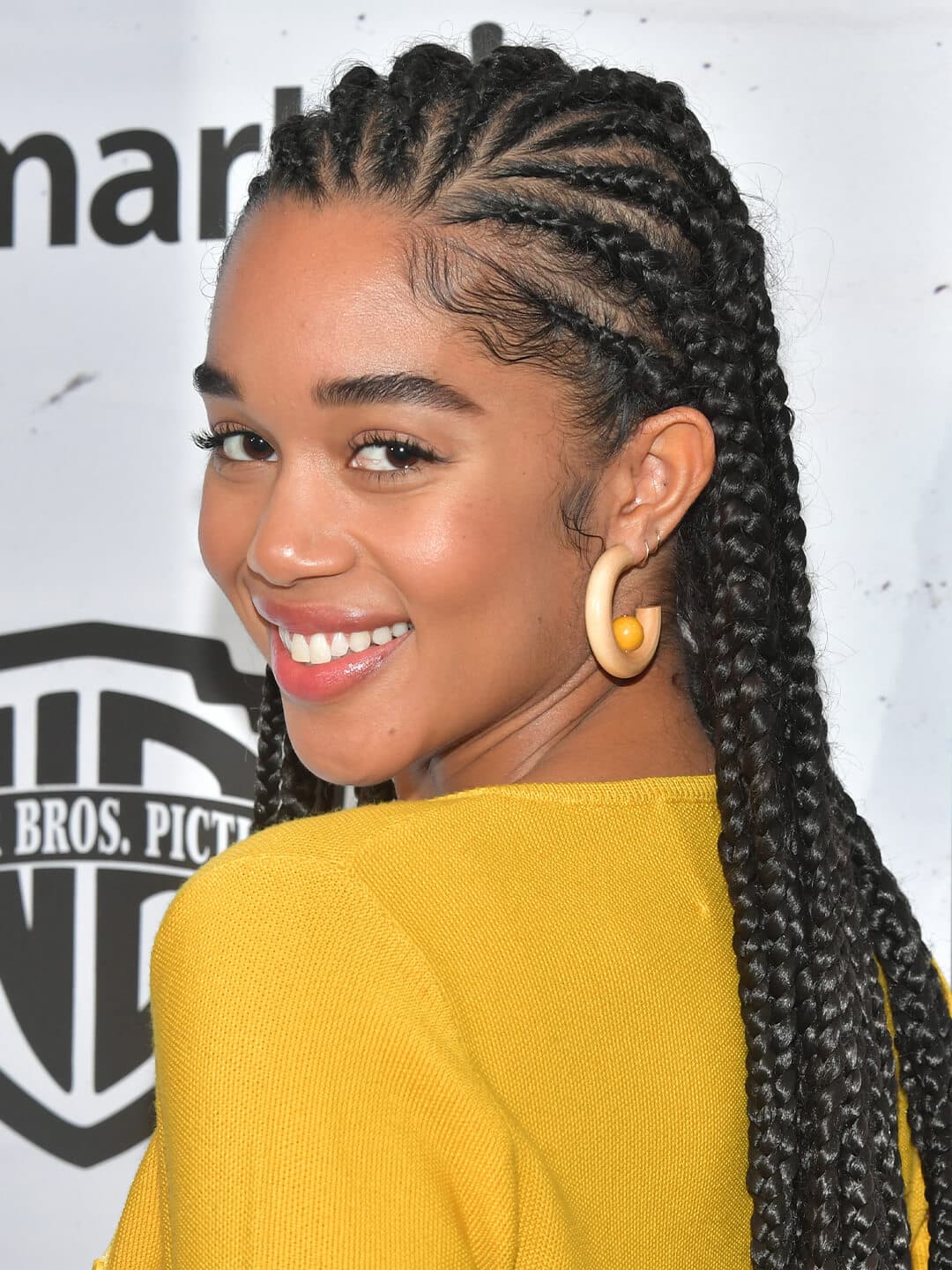 Smiling Laura Harrier wearing a yellow dress and hoop earrings, and rocking a long and , goddess braid hairstyle