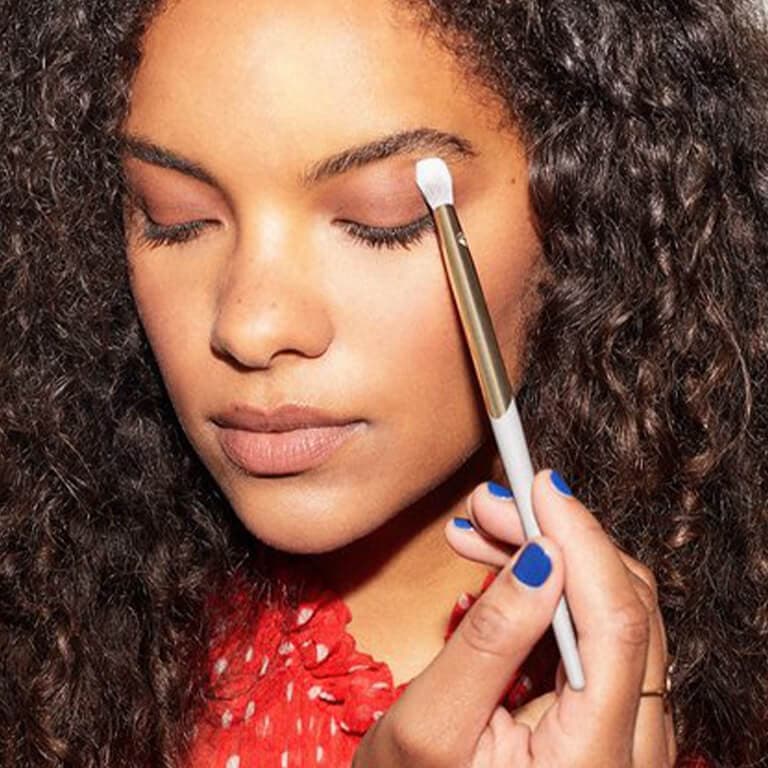 An image of a curly-haired model applying eye makeup on her crease using a COMPLEX CULTURE brush