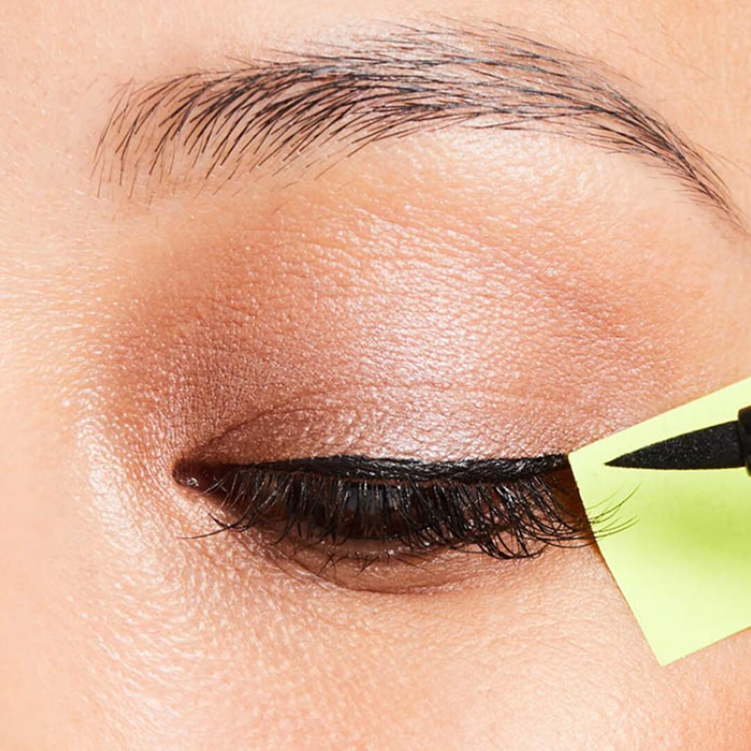 Close-up of woman's eyes, applying liquid eyeliner with sticky note as guide
