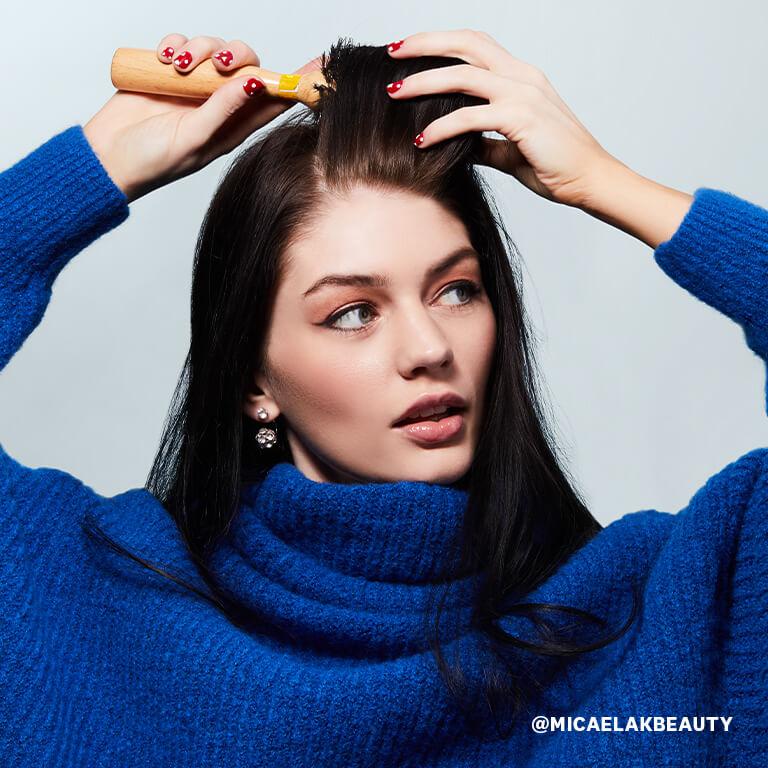 An image of a model with a shimmery pink eyeshadow wearing blue turtle neck knitted top and styling her hair with a brush