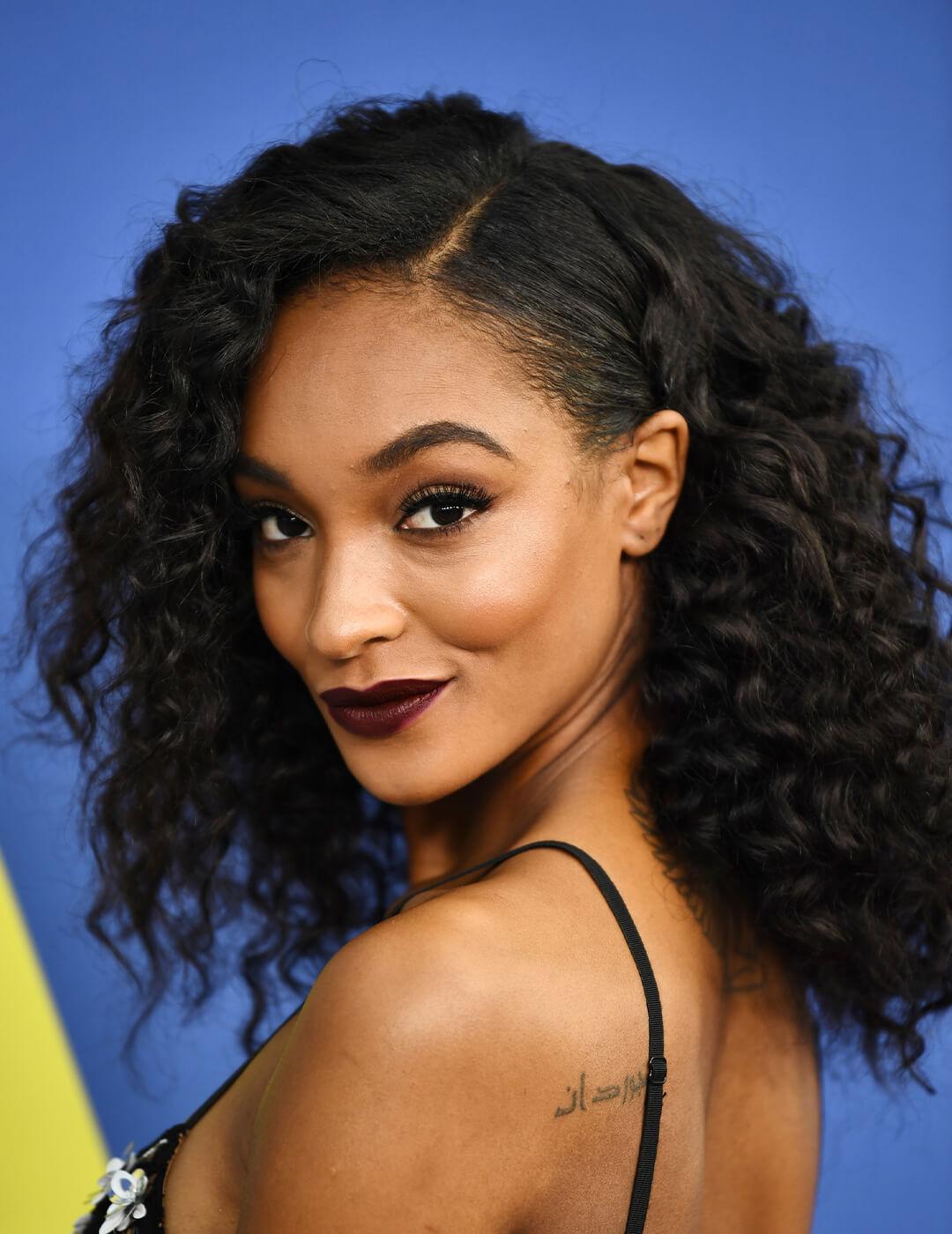 Jourdan Dunn in a black spaghetti strap dress rocking a parted, curly hairstyle while smirking