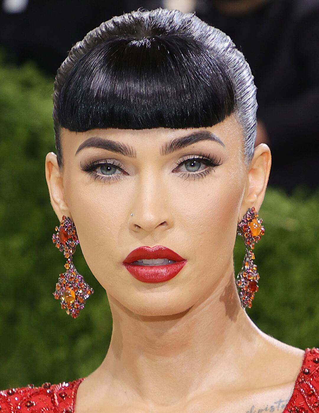 Megan Fox looking glam in a slick hairstyle with full bangs, red-stoned dangling earrings, and minimal makeup look paired with bright red lips
