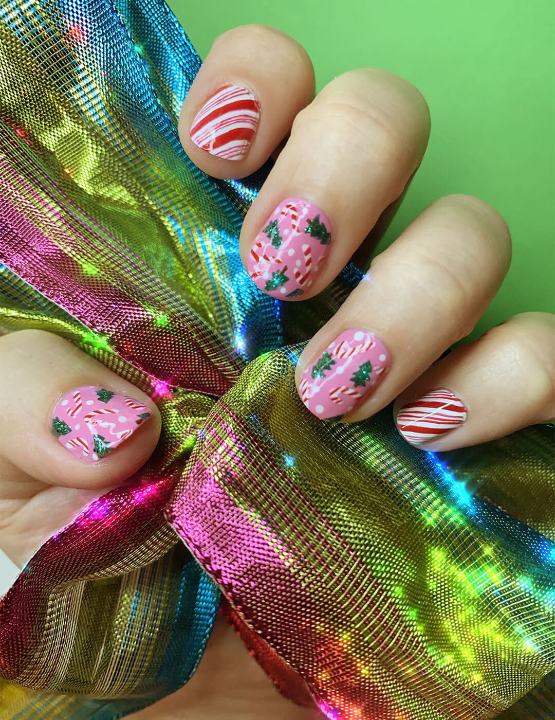 Close-up image of a model's hand with a peppermint, candy cane, and Christmas tree themed nail art mani holding a shiny rainbow colored ribbon