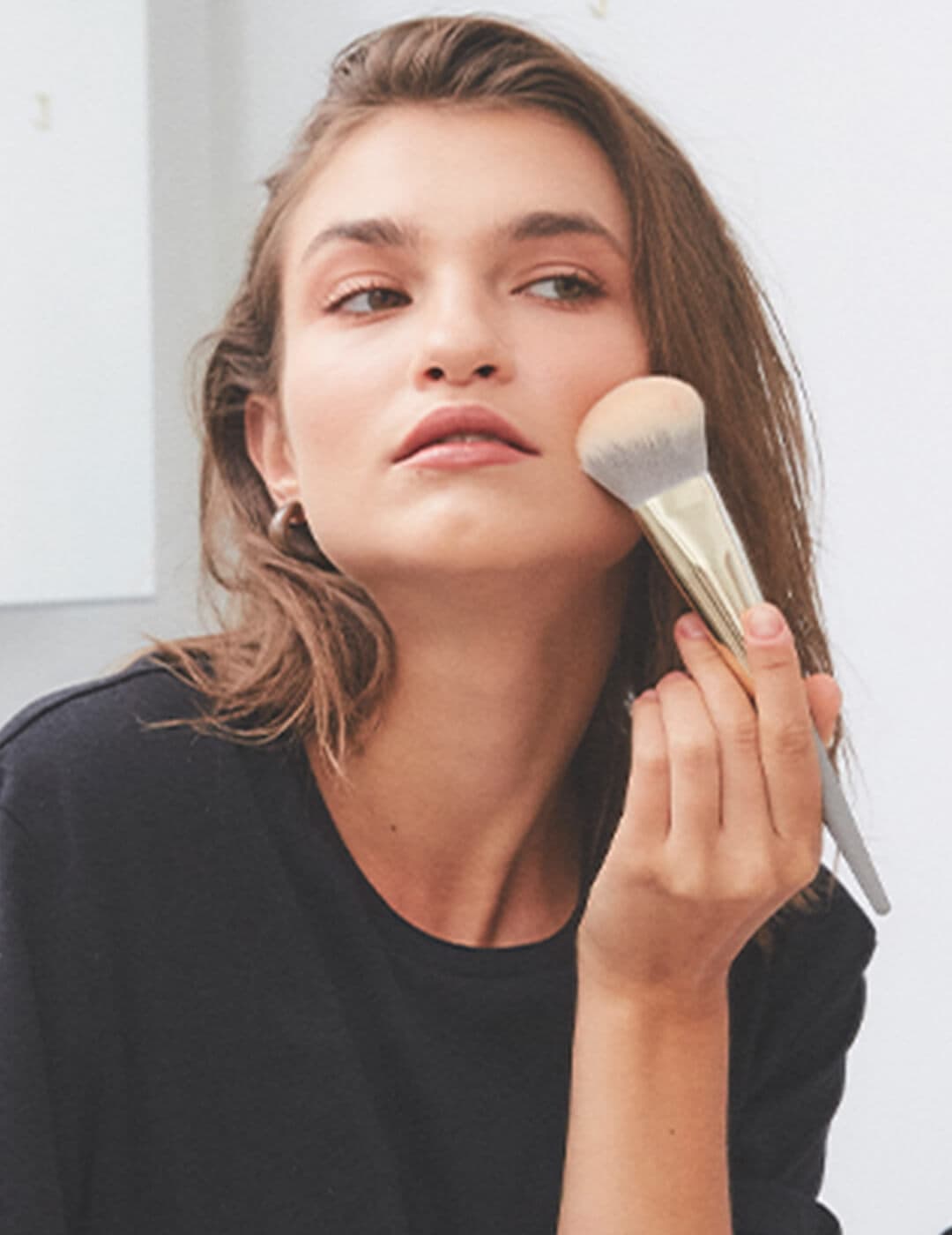 A photo of a woman wearing black top holding a powder brush pressed against her cheeks