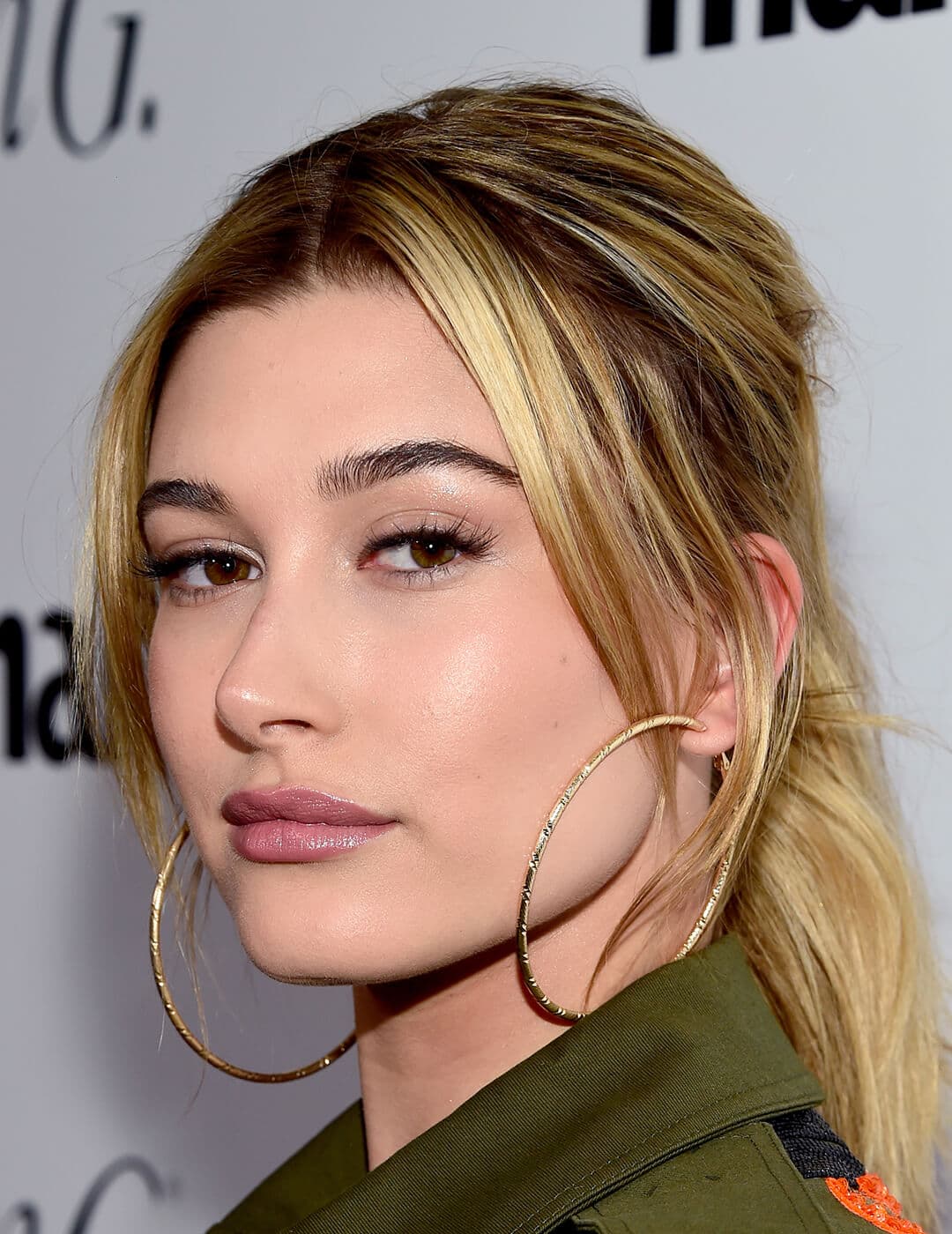 Close-up image of Hailey Bieber rocking an olive green jacket, large hoop earrings, and ponytail hairstyle with curtain bangs