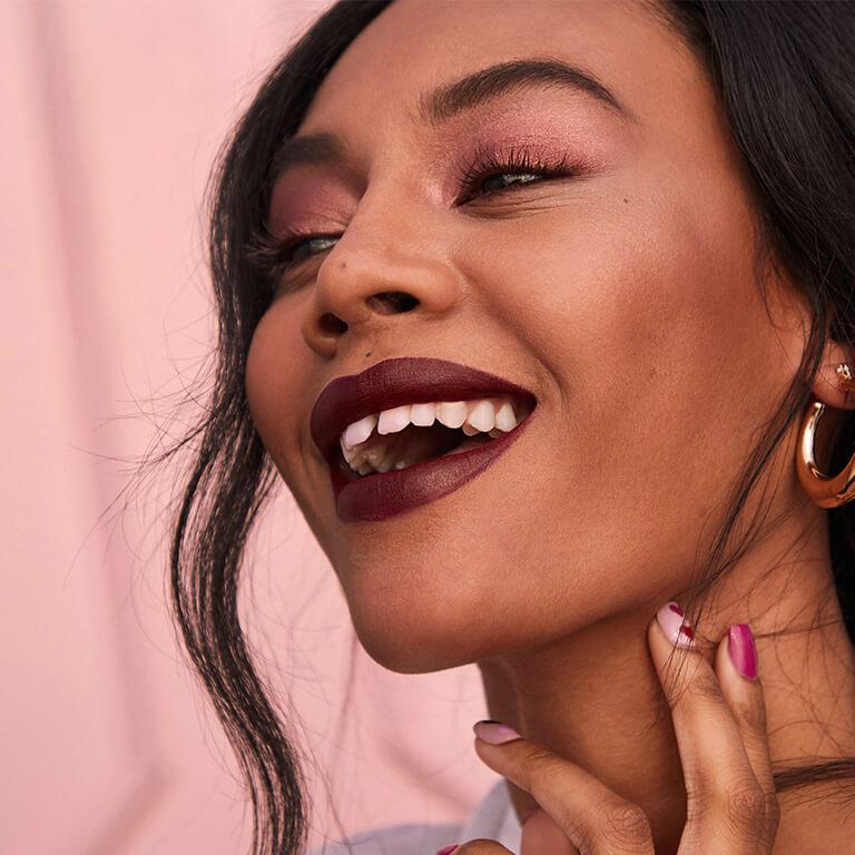 A closeup image of a model wearing rose gold eyeshadow and matte dark mauve lipstick and gold hoop earrings