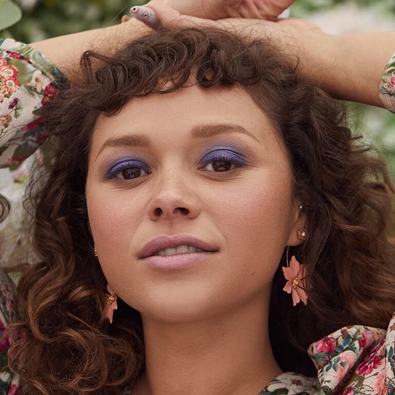 A close-up image of a curly-haired model rocking a dark blue smoky eyeshadow look and wearing floral earrings