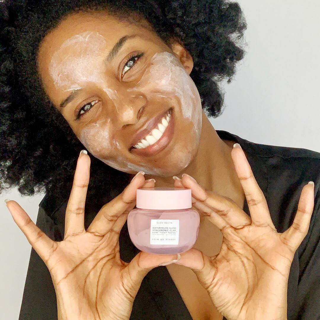 Close-up of a black woman holding the GLOW RECIPE Watermelon Glow Hyaluronic Clay Pore-Tight Facial with mask on her face