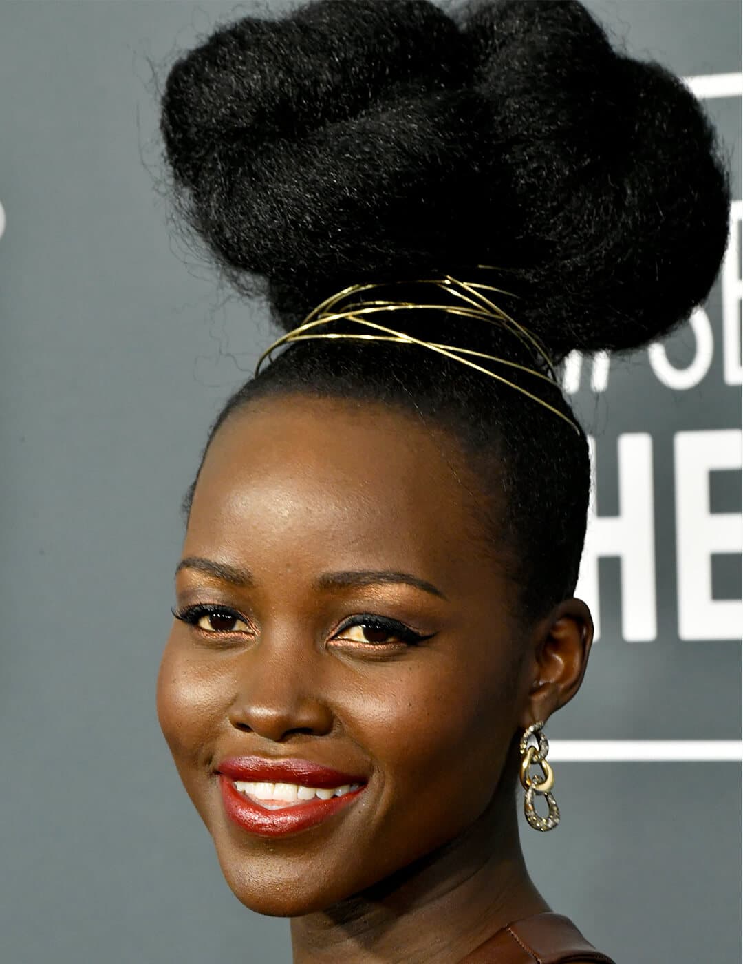 Close-up of Lupita Nyong'o rocking a high bun hairstyle wrapped with gold twine, copper eyeshadow makeup look paired with a berry lipstick, and gold links earrings