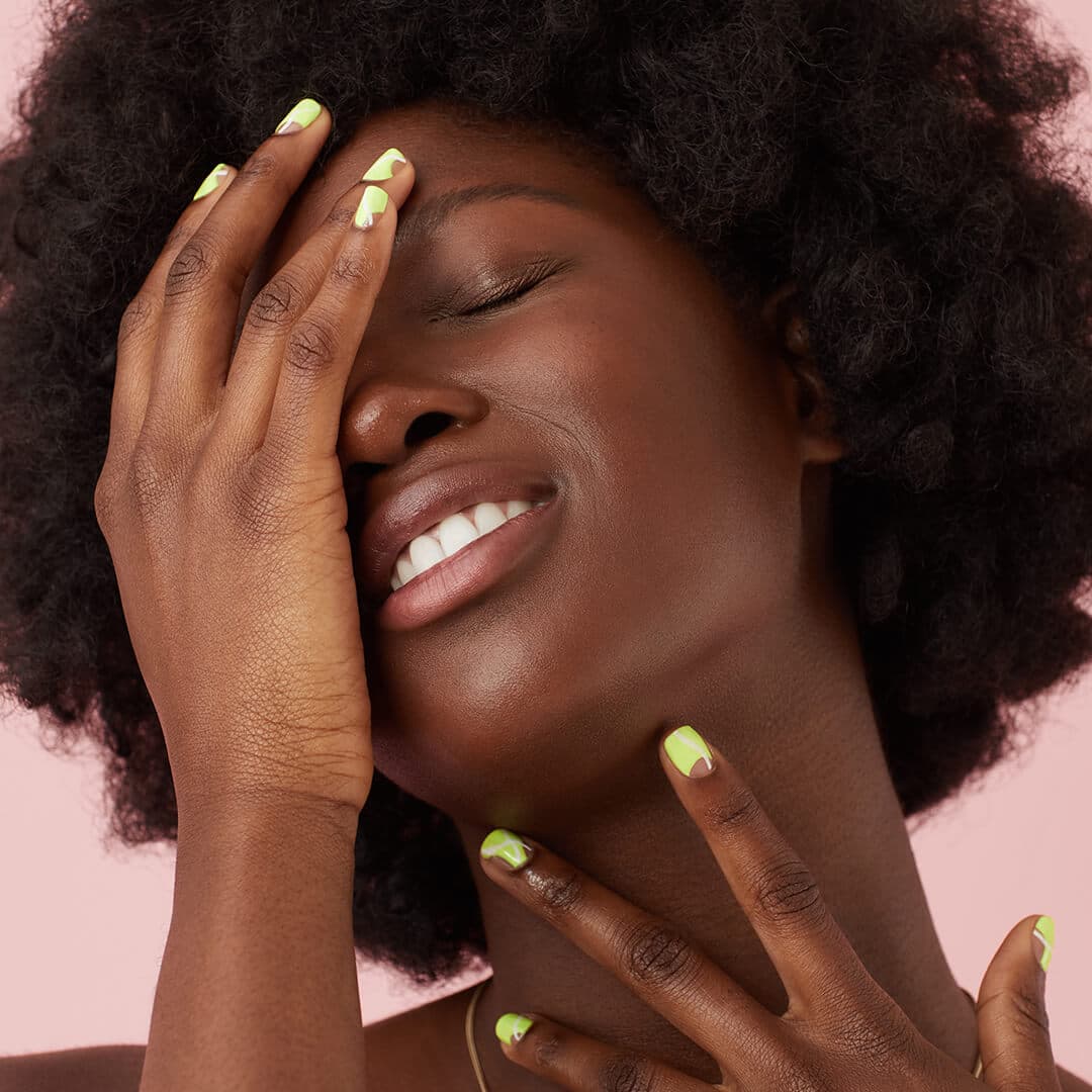 Close-up of a black model with slime green nail art posing with eyes closed against pink background