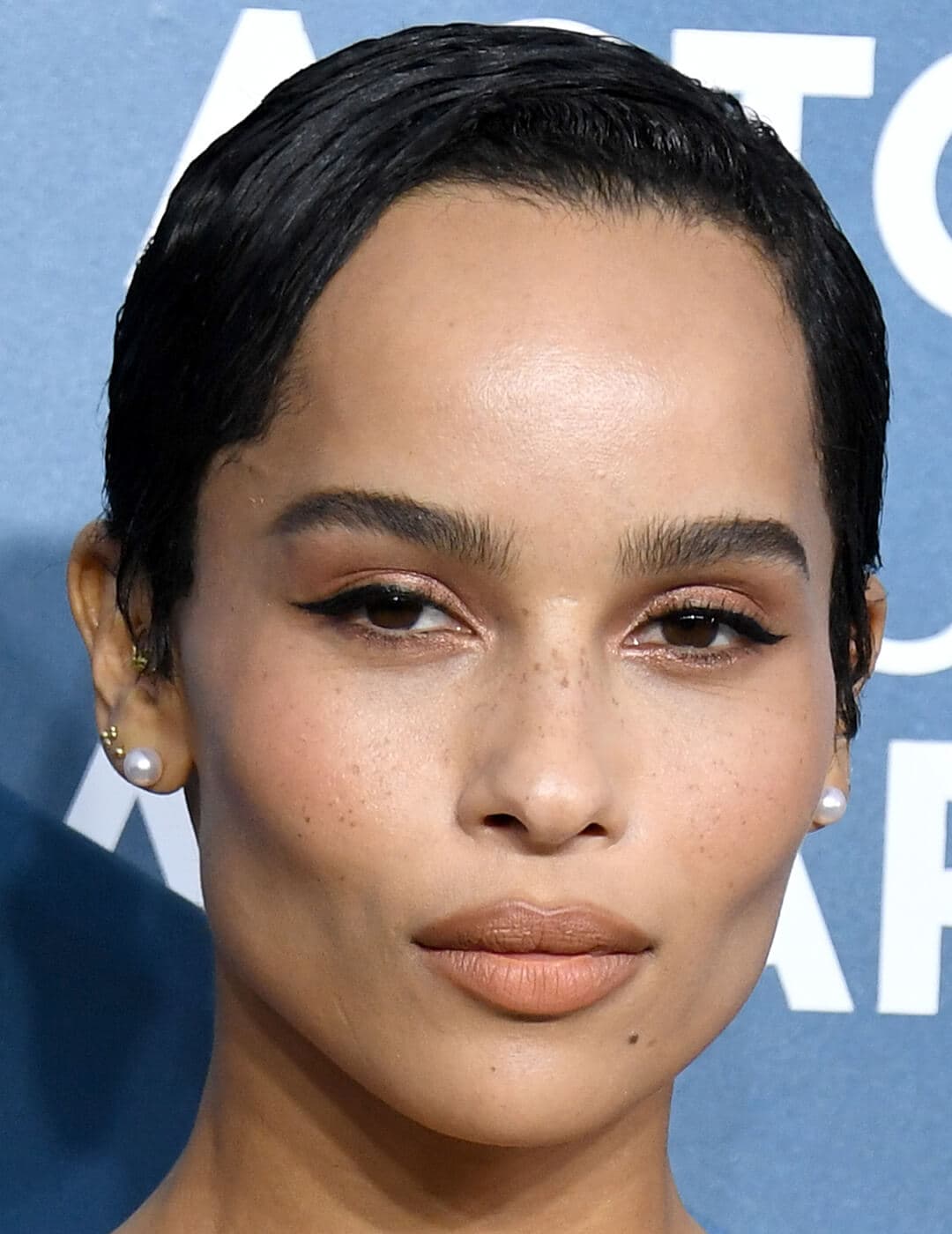 A closeup of Zoë Kravitz with pixie cut hairstyle, wearing black eyeliner and shimmery brown eyeshadow with a nude lipstick color 