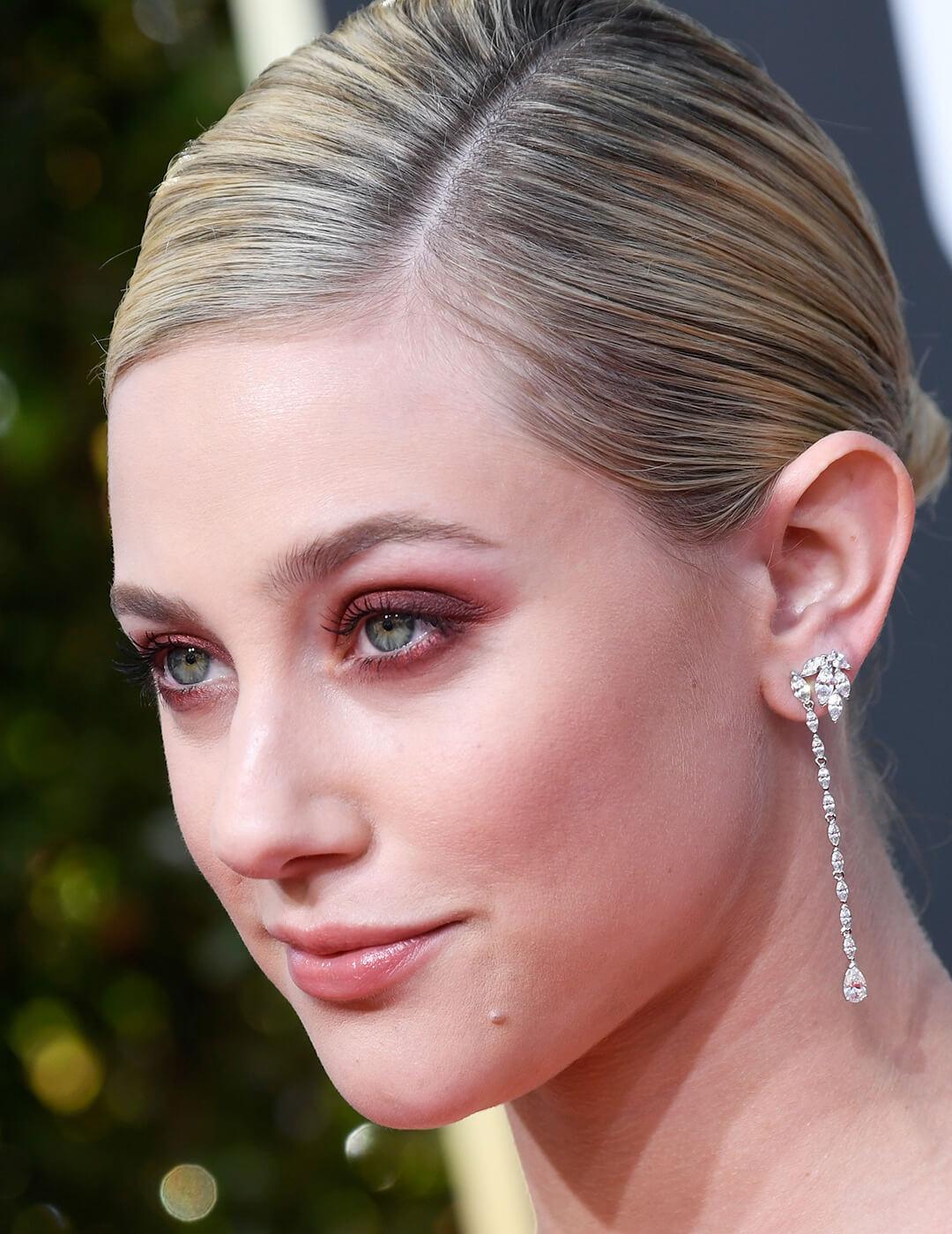 Close-up of Lili Reinhart rocking a slick back hairstyle, soft red smoky eye makeup, and silver dangling earrings