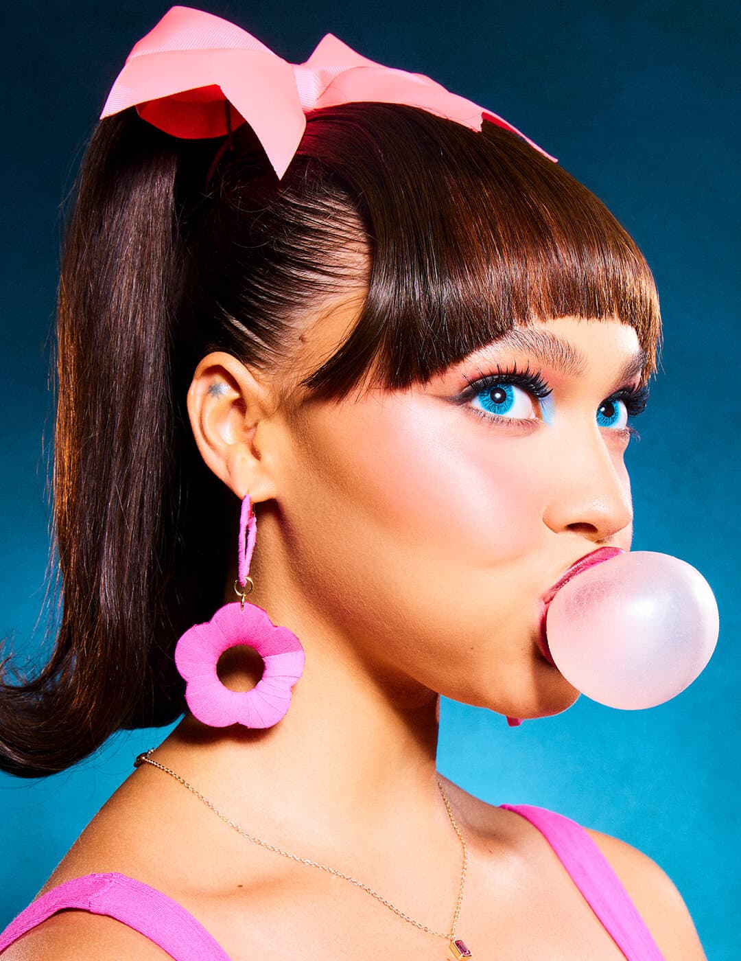 Model in a blue eye makeup look, pink dress, earrings, and bow blowing a bubblegum