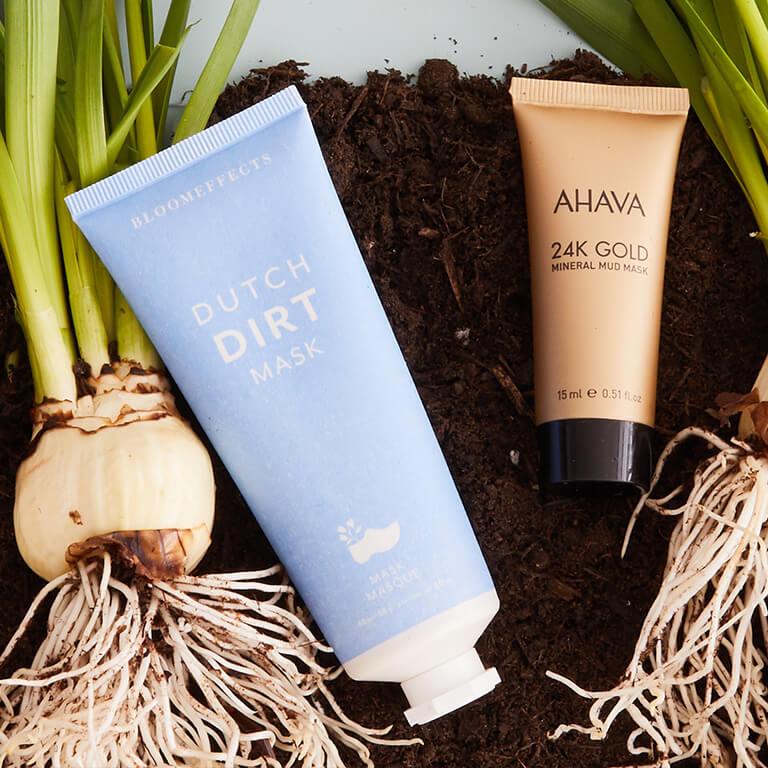 A flatlay image of BLOOMEFFECTS Dutch Dirt Mask and AHAVA 24K Gold Mineral Mud Mask and orchid bulbs on dirt soil