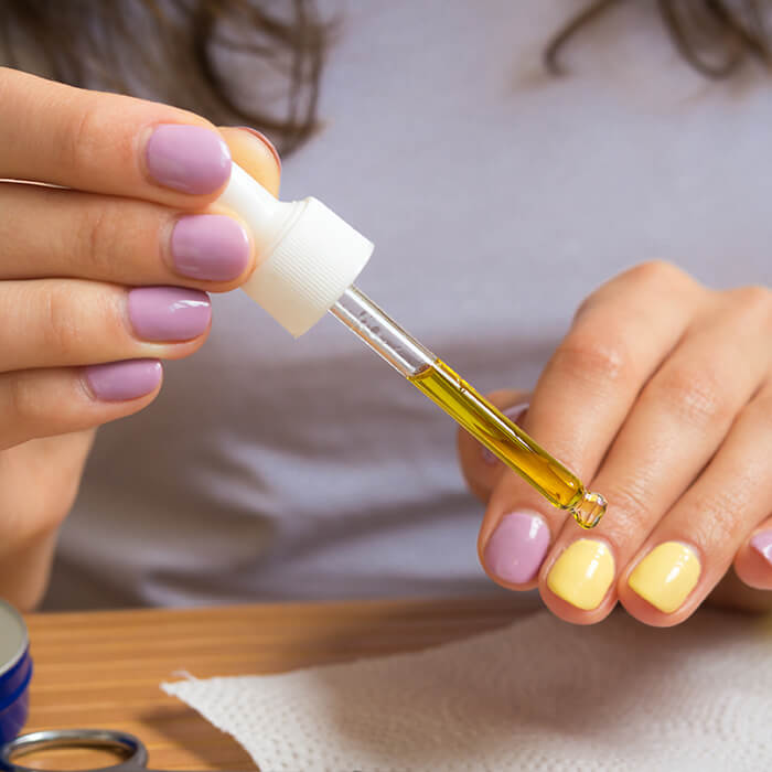 Close-up of a woman's hands with lavender and yellow nail polish applying oil on her cuticles with a dropper