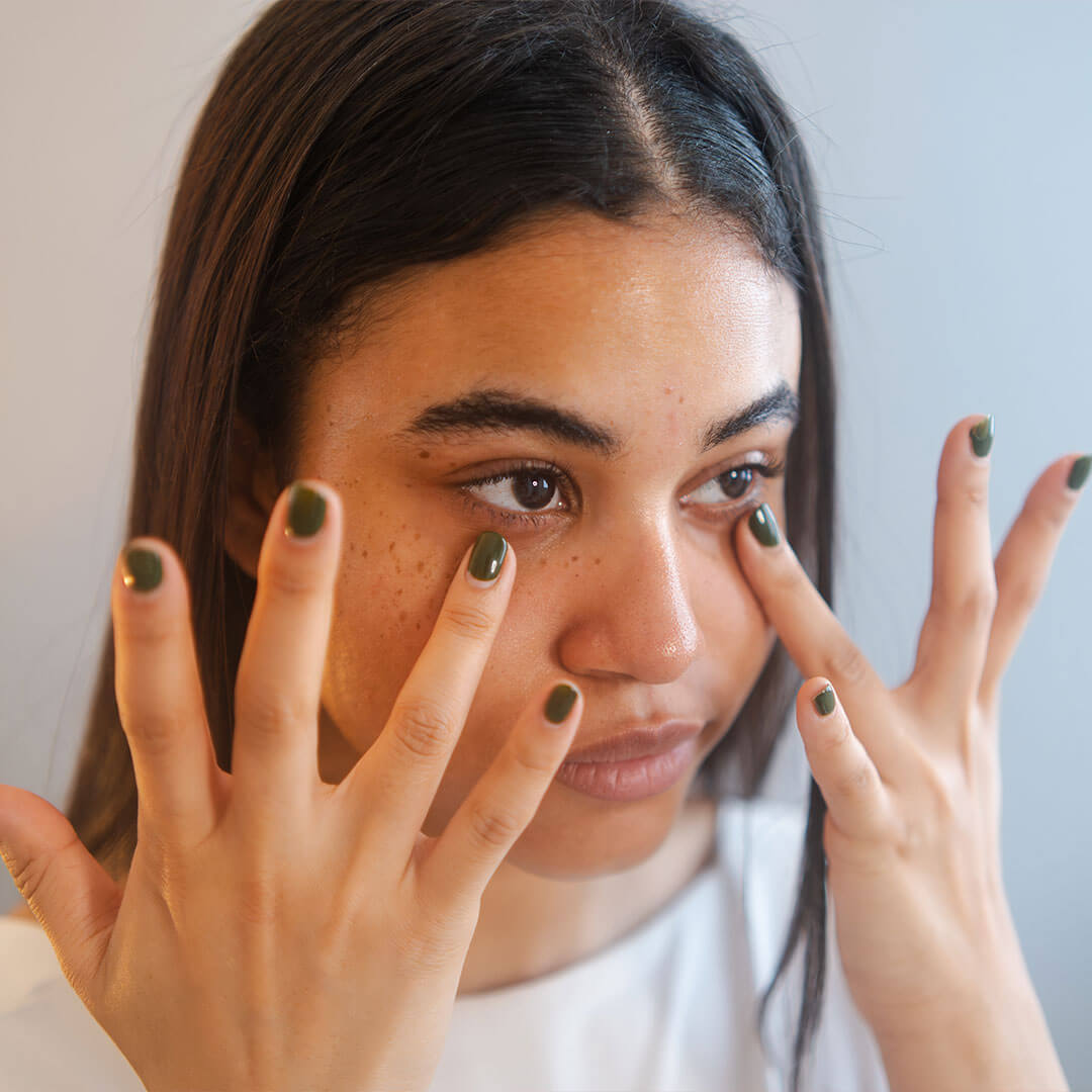 A close-up shot of a woman with black nails applying serum to her under-eye area