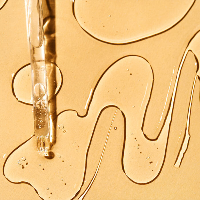 A picture of a dropper with serum drops splattered against a soft yellow backdrop