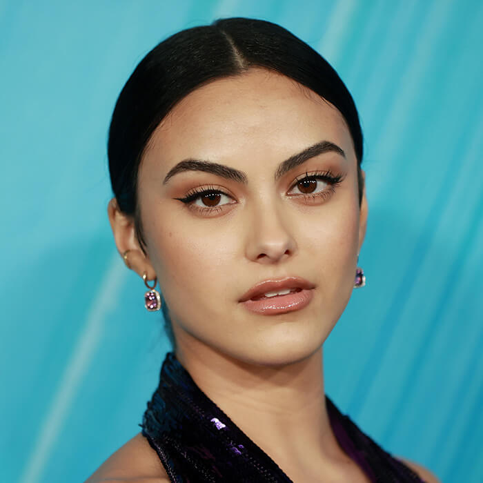 An image of Camila Mendes with her sleek blue-black hair, gazing at the camera, wearing black eyeliner and a stylish dark halter top purple dress
