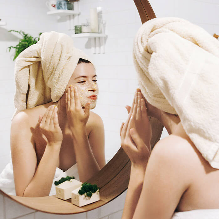 Woman in a towel scrubbing her face while looking in the mirror of her bathroom