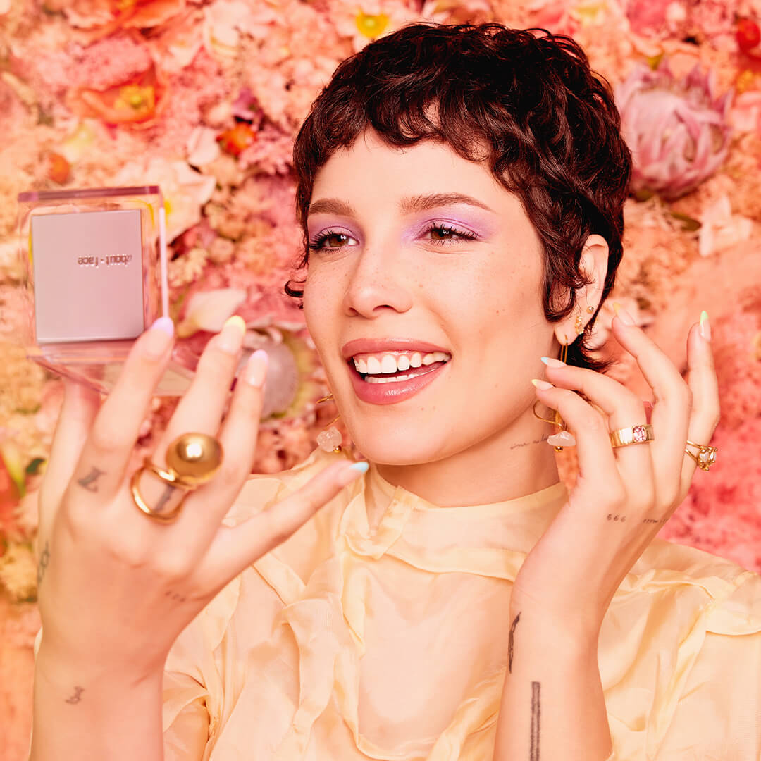 Close-up of a smiling Halsey looking at herself in a compact mirror against floral background