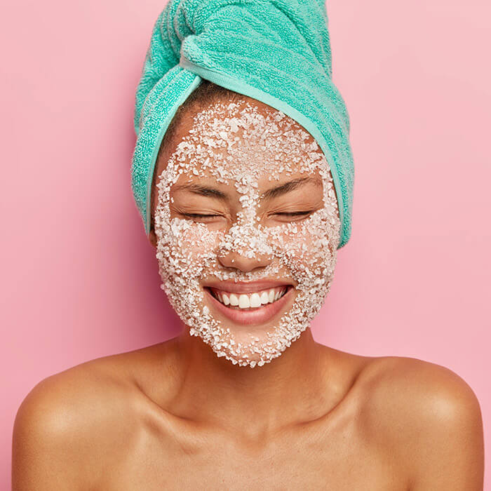 Close-up of a woman with a green towel around her hair and facial scrub on her face smiling with eyes closed on pink background