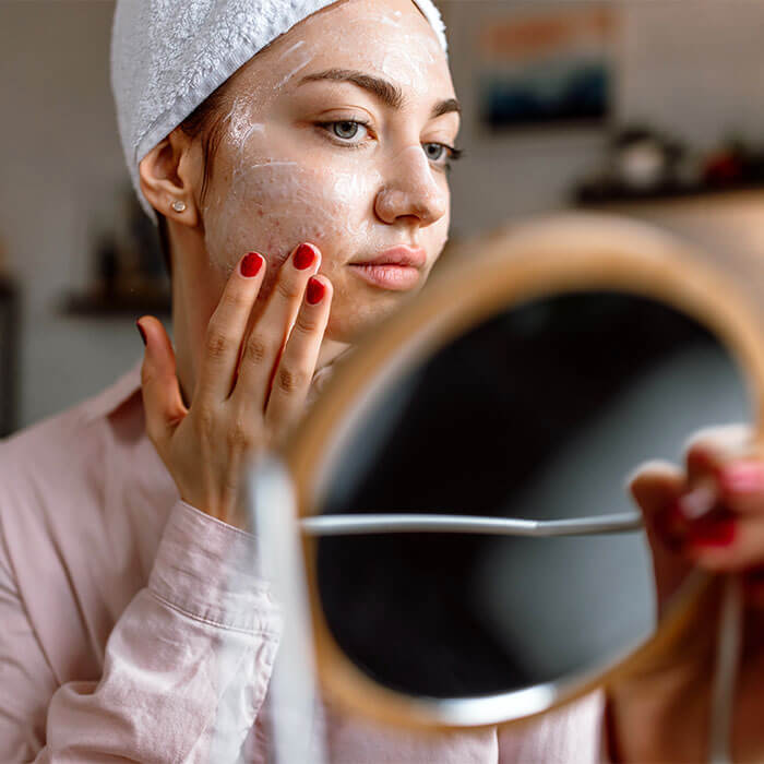An image of a young woman with acne skin applying cream on her face while holding a mirror