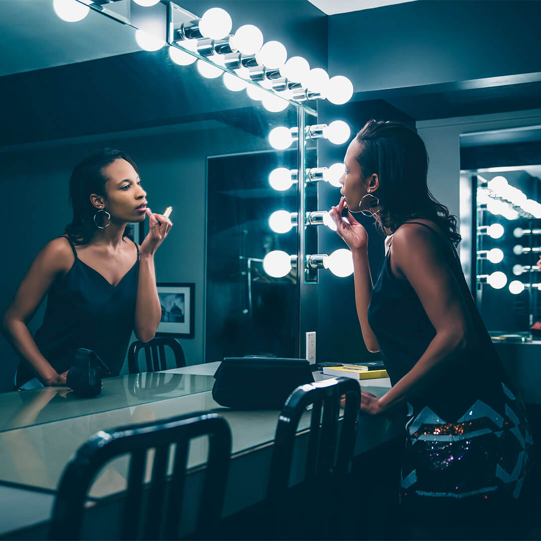 An image of a woman of color applying lipstick  in front of a brightly lit vanity mirror, dressed in a black satin tank top and sporting a hoop earring