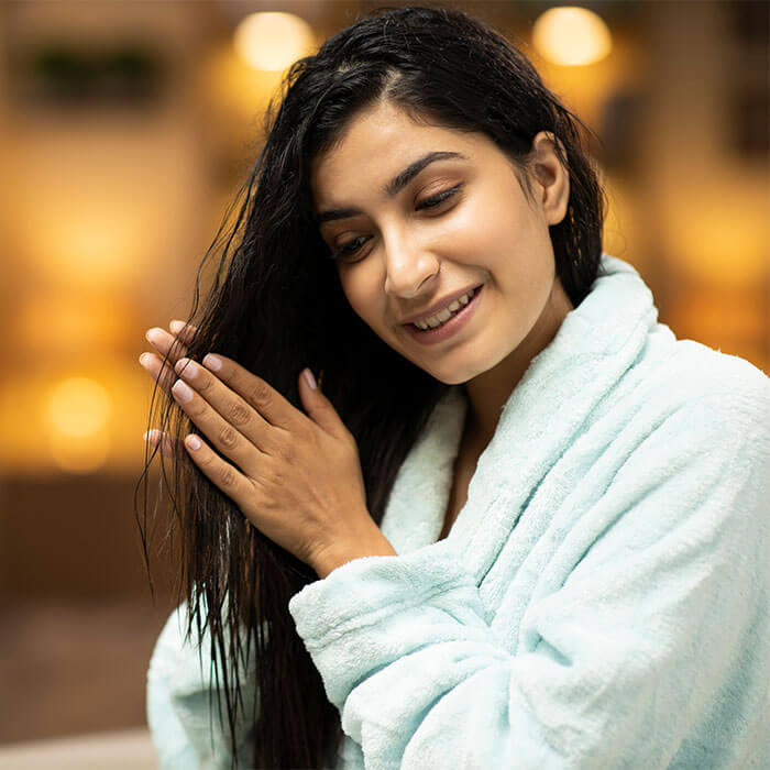 A picture shows a woman wearing a light blue bathrobe and oiling her hair after getting out of the shower