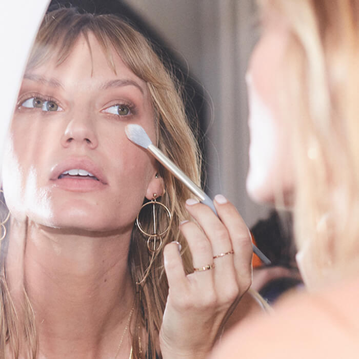 A photo of a woman looking at herself in the mirror while applying cosmetics on her face