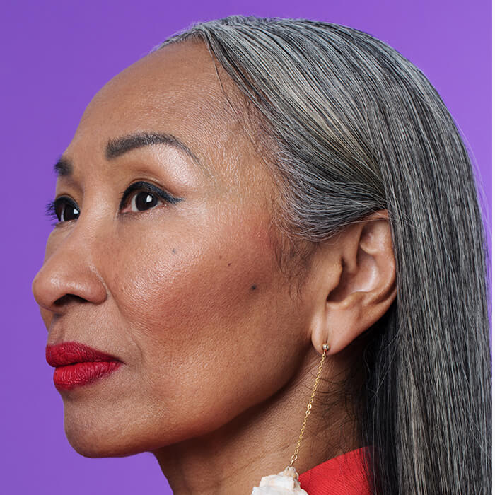 A closeup photo of a mature woman wearing a red coat and conch earrings on a purple background