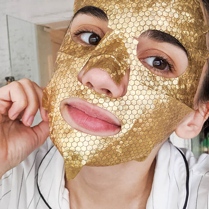 Photo of a young woman in her apartment putting on a gold sheet mask