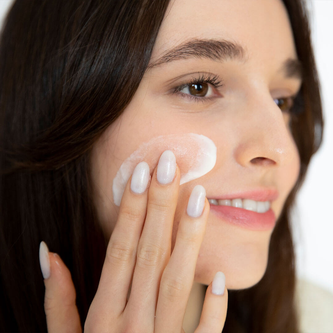 An image of a woman with clear skin, her nails manicured in a white nail polish applying a moisturizer while smiling sideways