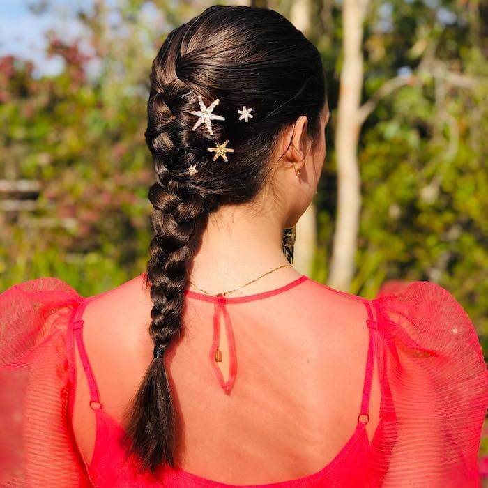An image of a woman with her back to the camera showcasing her long braided black hair adorned with star-shaped hairpins dressed in a red, sheer top