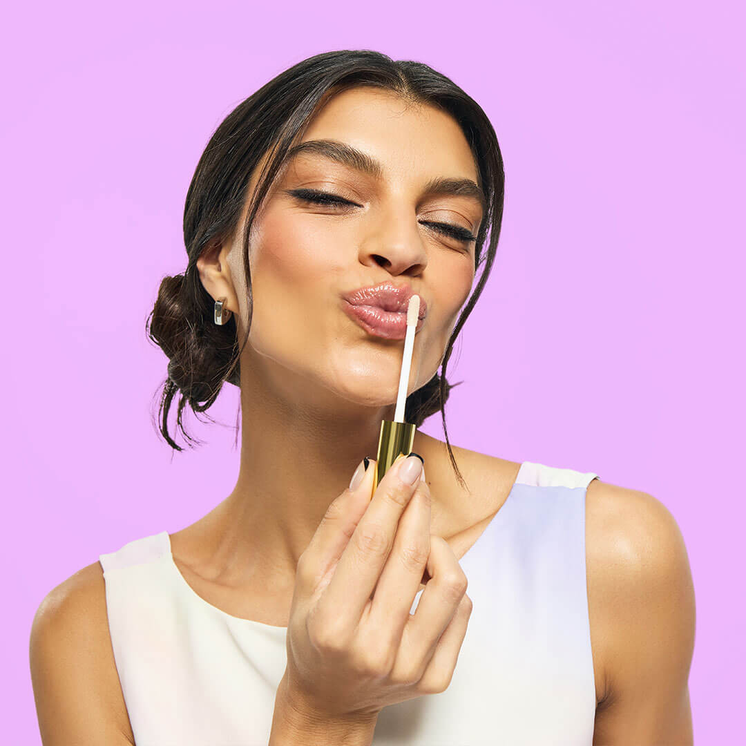 A woman pouts playfully while applying a shiny lip product with a gold applicator against a vibrant lavender background