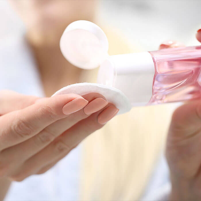A close-up shot of hands carefully pouring a toner or serum from a clear pink bottle onto a cotton pad